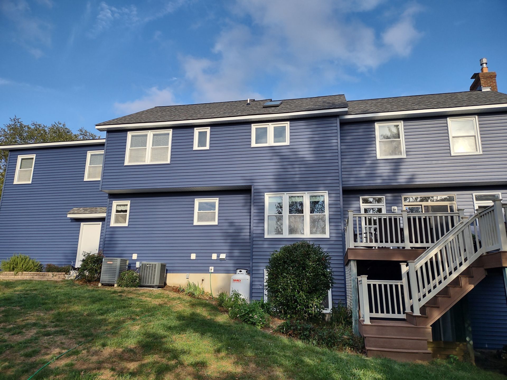 Blue-sided house with a dark roof, a deck, and air conditioning units, set against a blue sky.