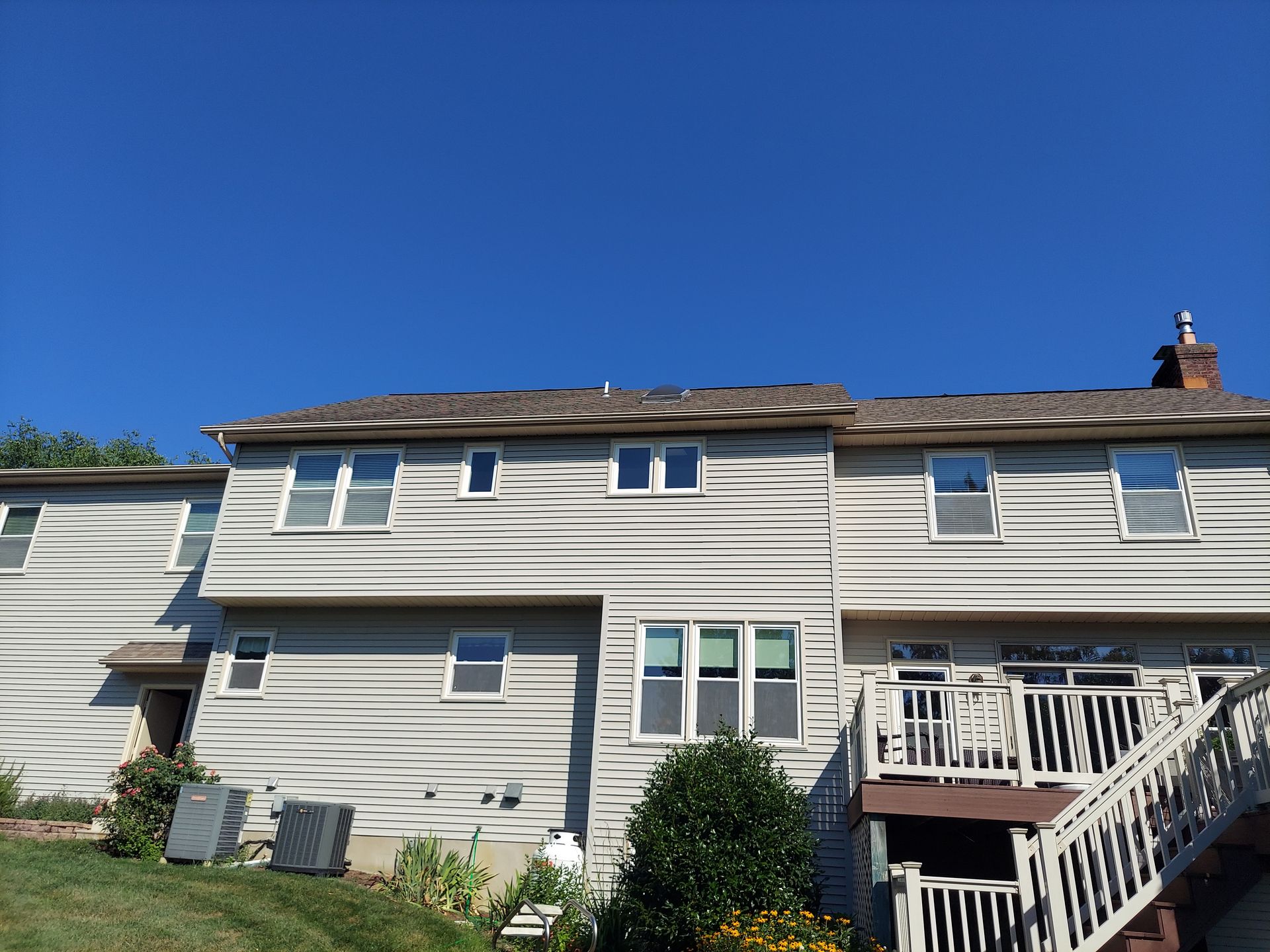 Back of a two-story house with tan siding, windows, and a deck, under a clear blue sky.