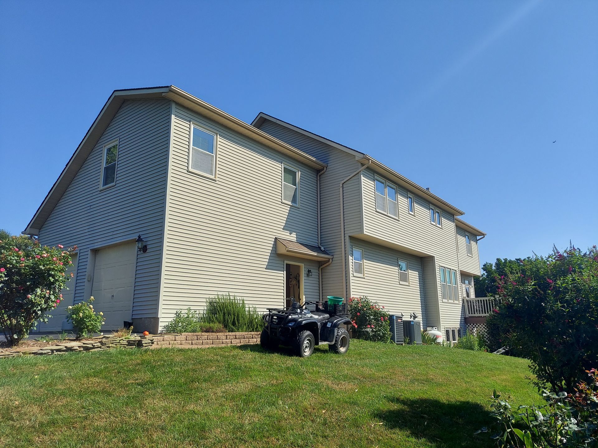 Two-story house with tan siding, a garage, and an ATV parked on a grassy hill under a blue sky.