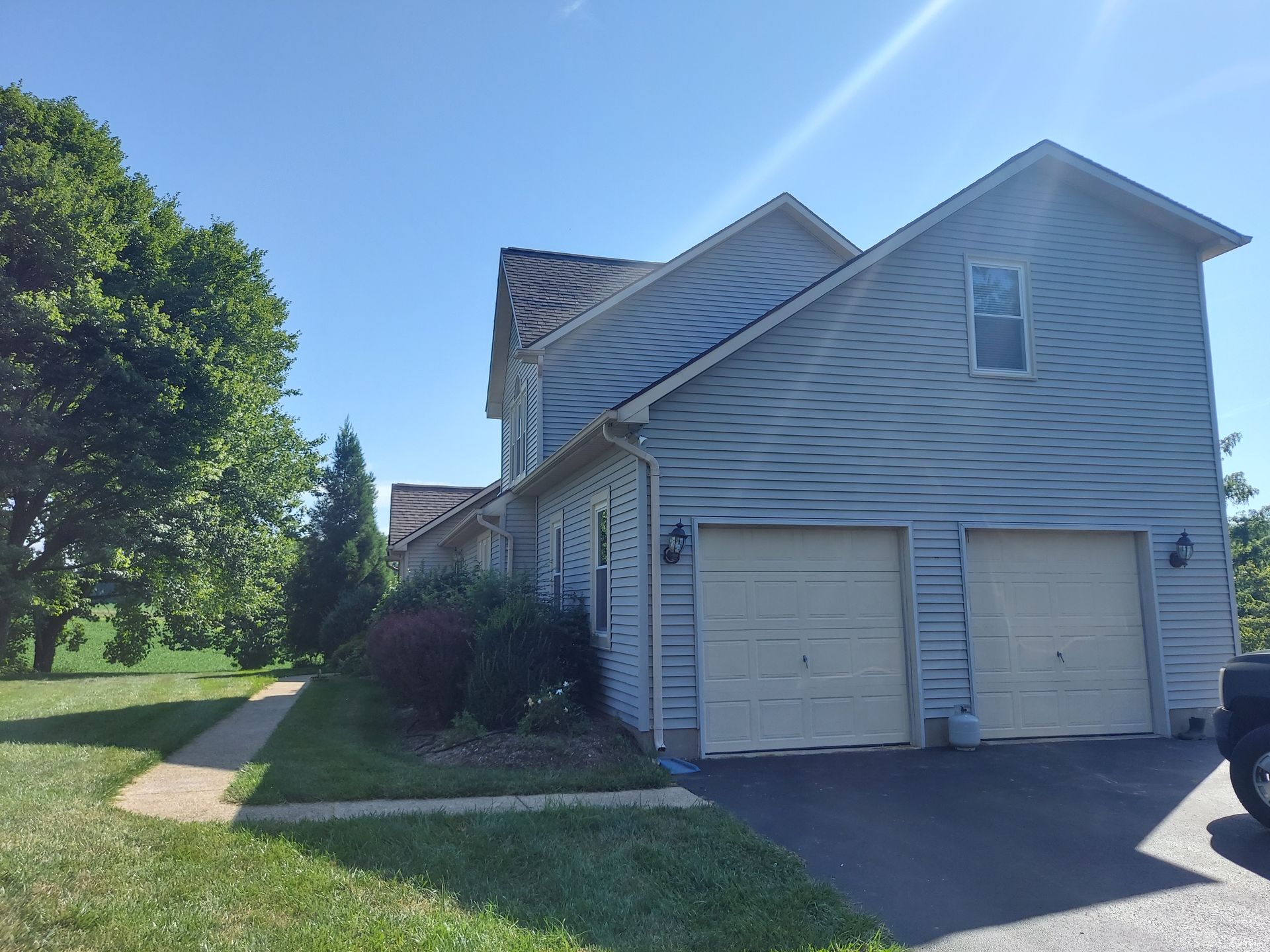 Two-story house with blue siding, two-car garage, and a paved driveway. Green lawn and trees surround it.