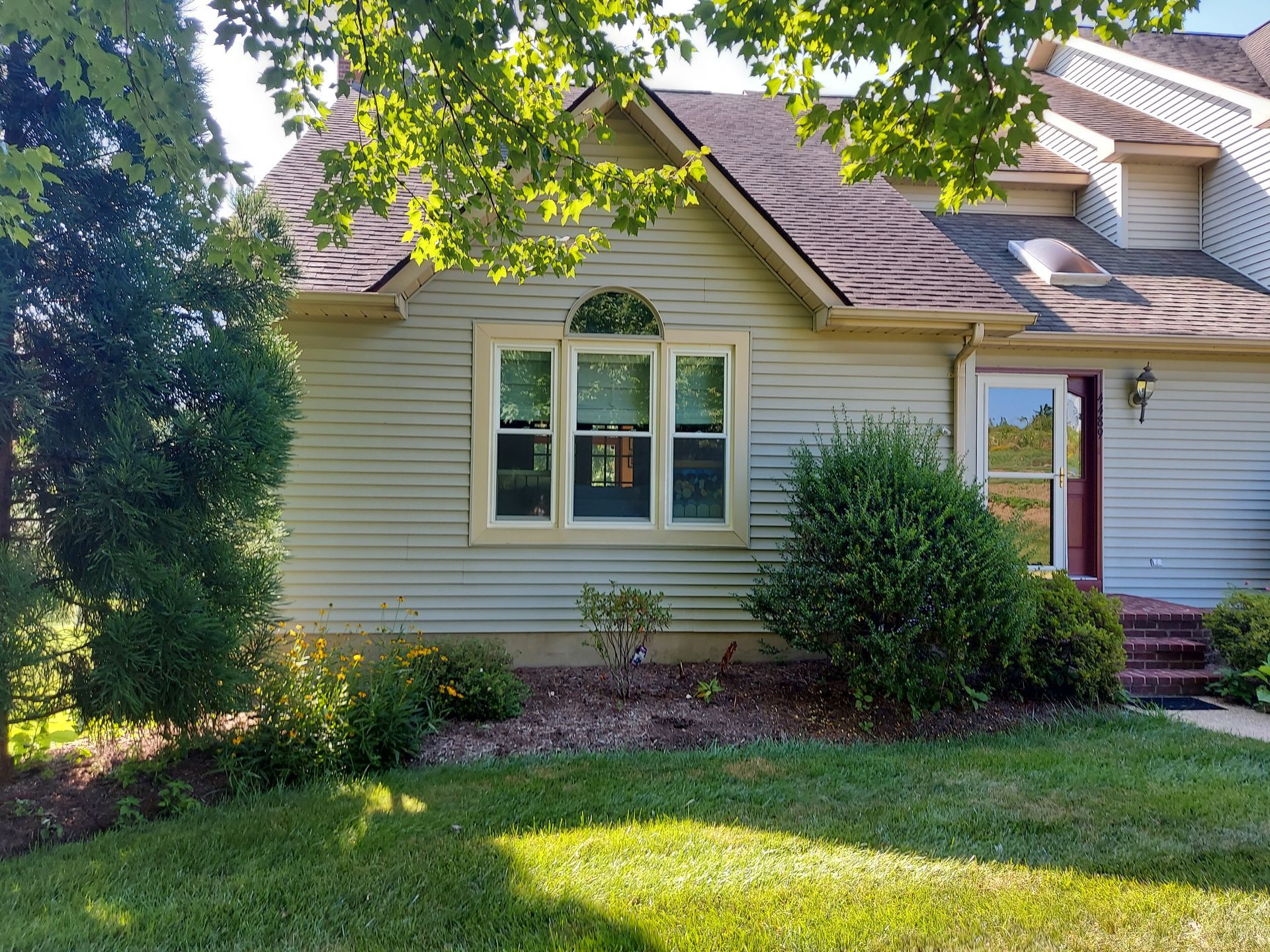 A light green house with a brown roof and window, a front yard of grass and bushes.