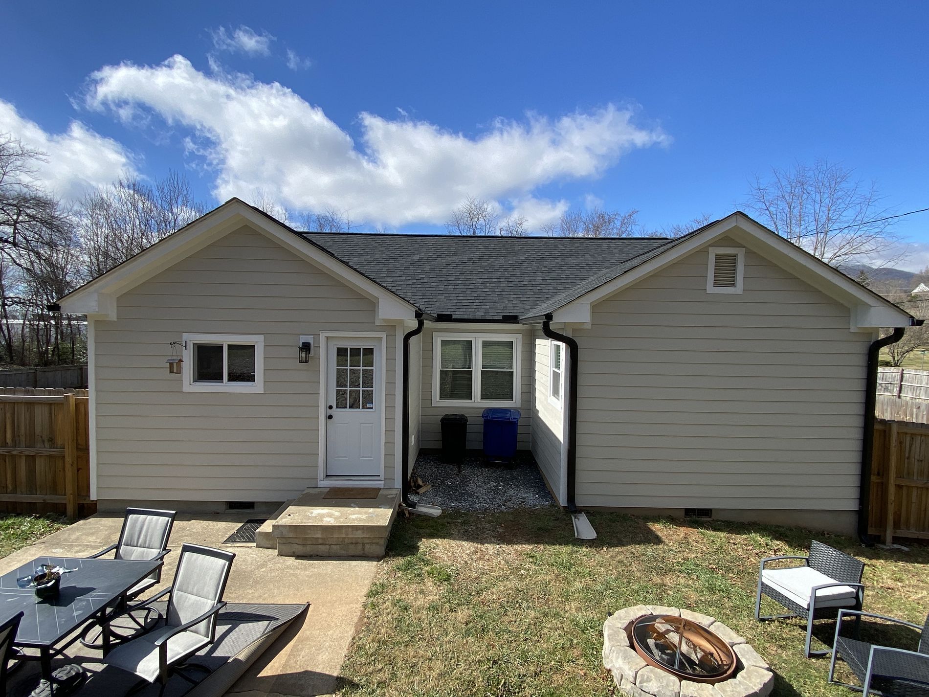 Backyard of a beige house with a patio, chairs, and a fire pit under a blue sky.