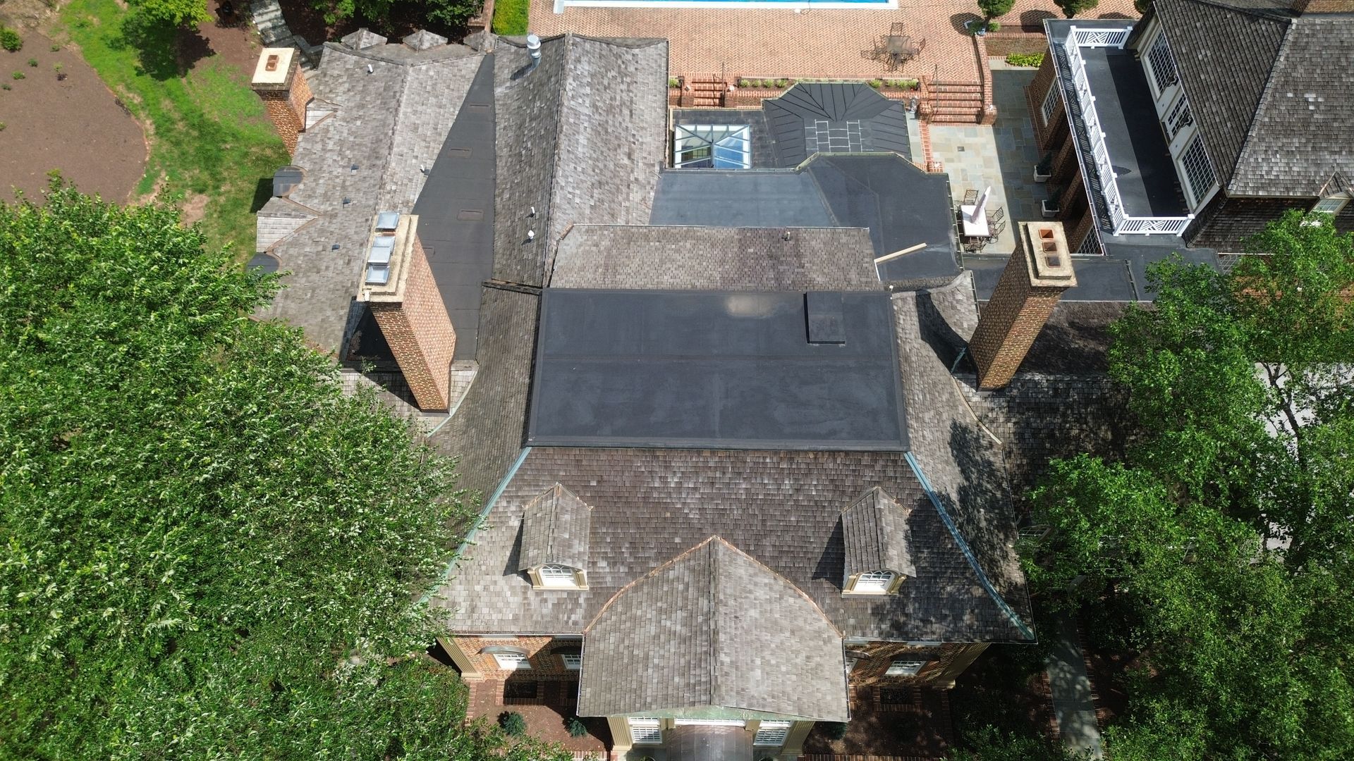 Overhead view of a multi-level roof with chimneys, a pool, and trees in the background.