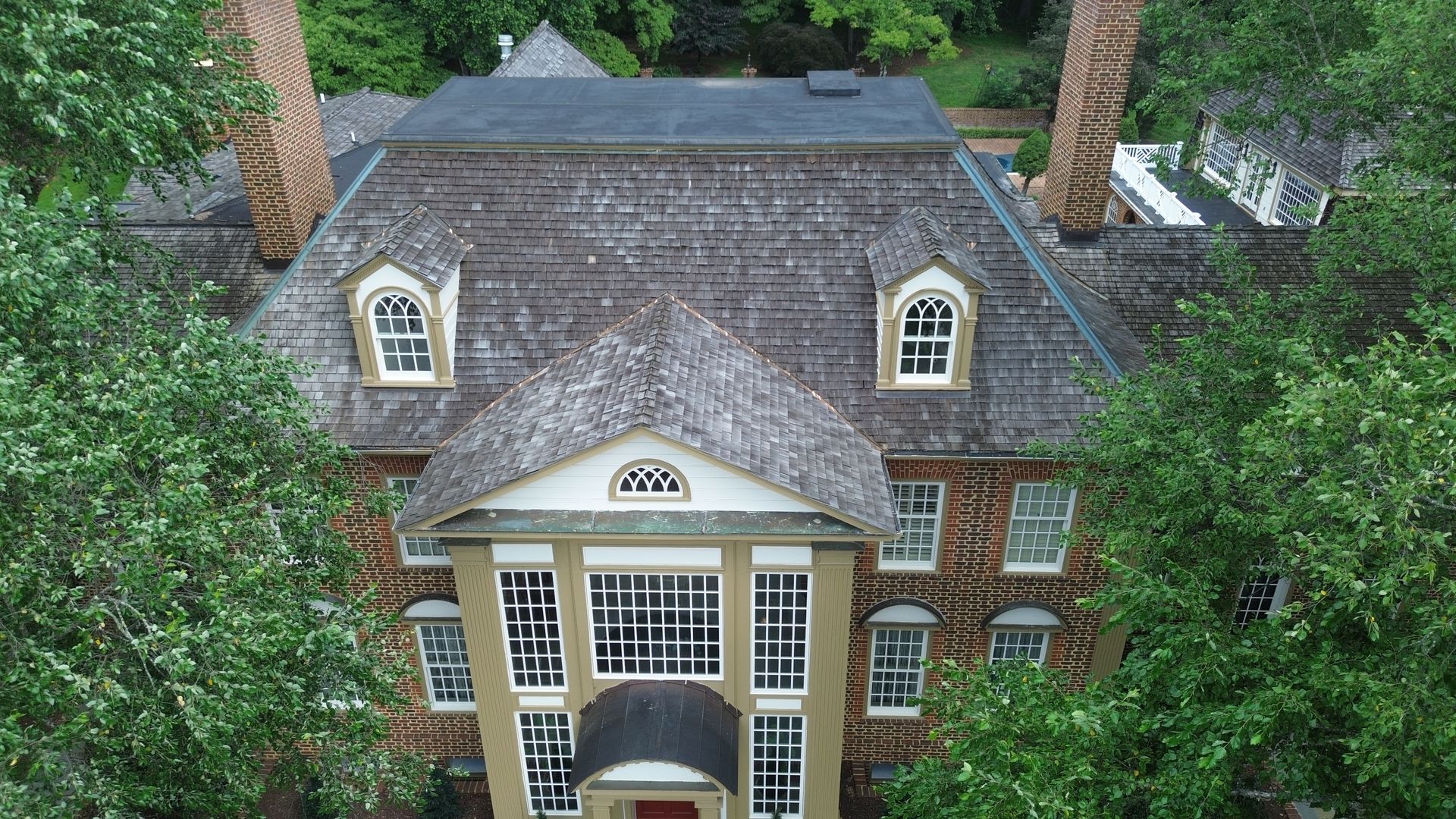 Brick mansion with gray roof, dormer windows, and white columns, framed by green trees.