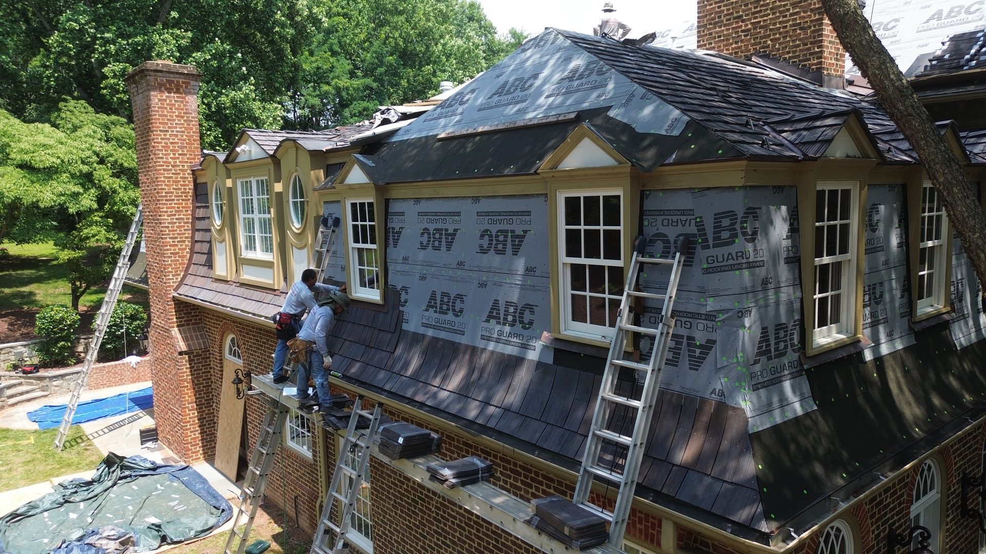 Workers installing roofing on a two-story brick house with dormer windows. Ladders and tools visible.