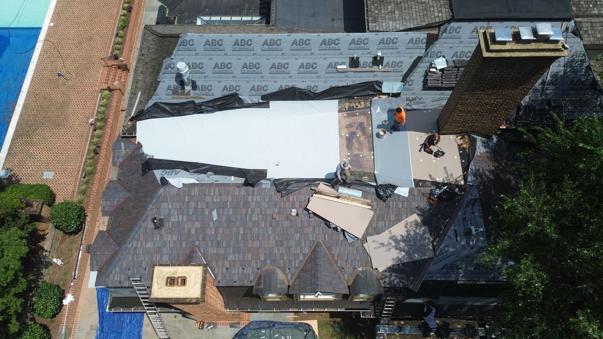 Overhead view of a house roof under construction. Workers on partially covered roof, blue tarp and pool visible.