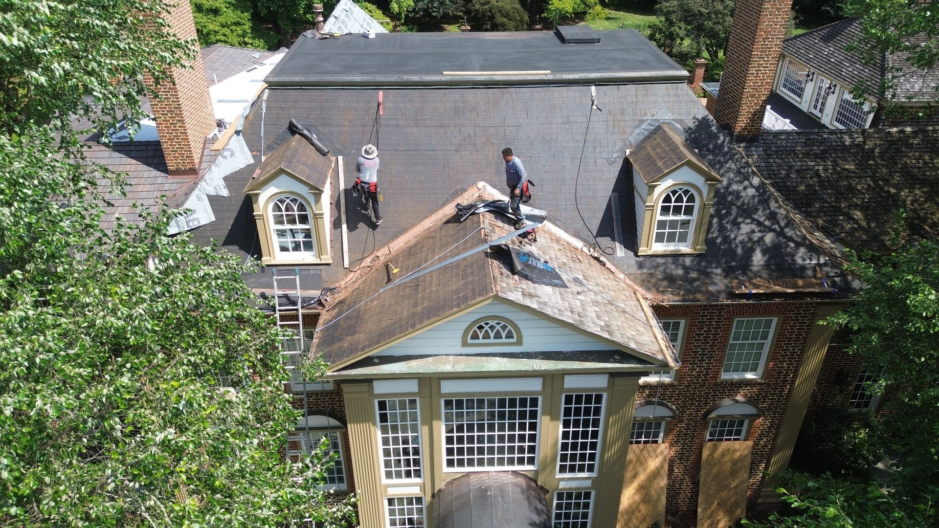 Workers on a roof, repairing a large brick and white building with windows and dormers.