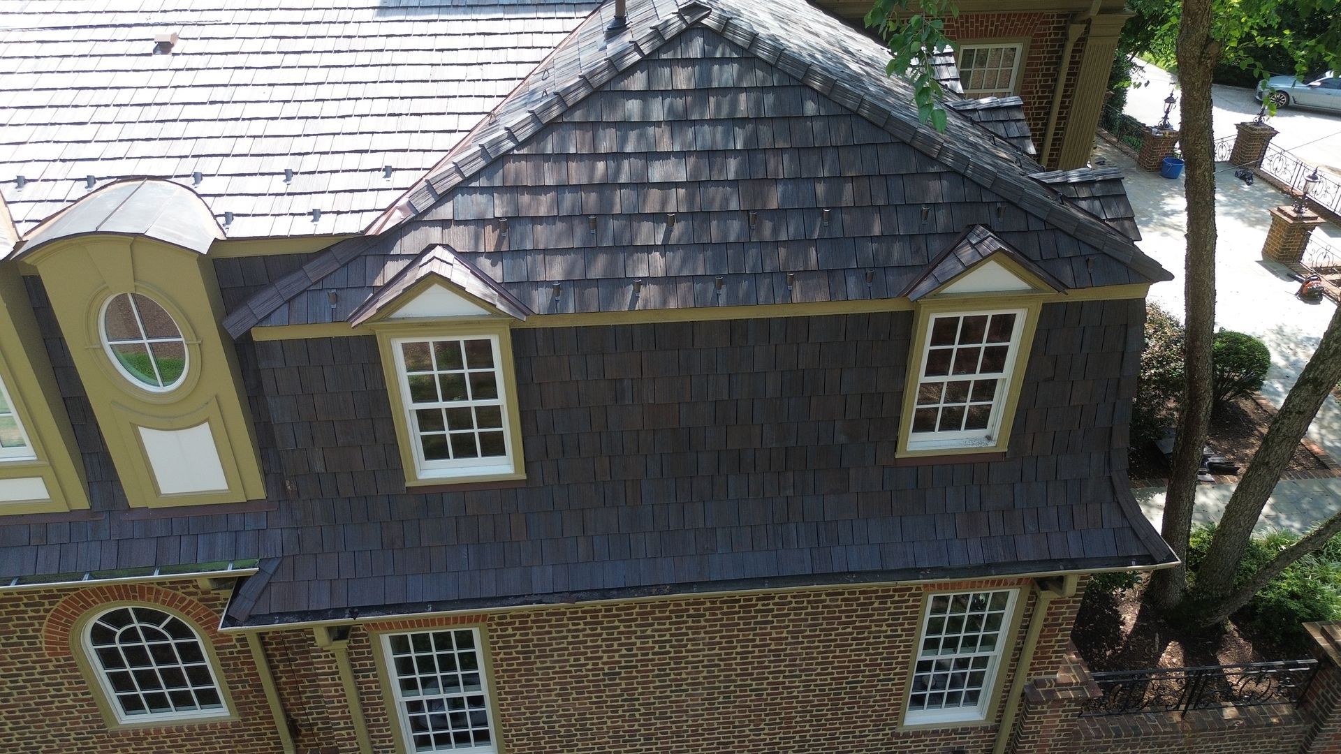 House with brick facade, dark roof, and multiple white-framed windows. A tree is visible on the right side.