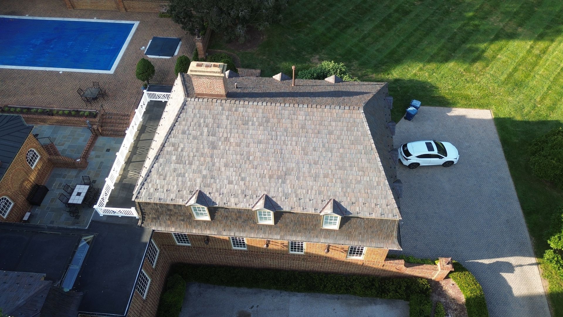Aerial view of a brick house with a gray roof, pool, and white car parked on the driveway.