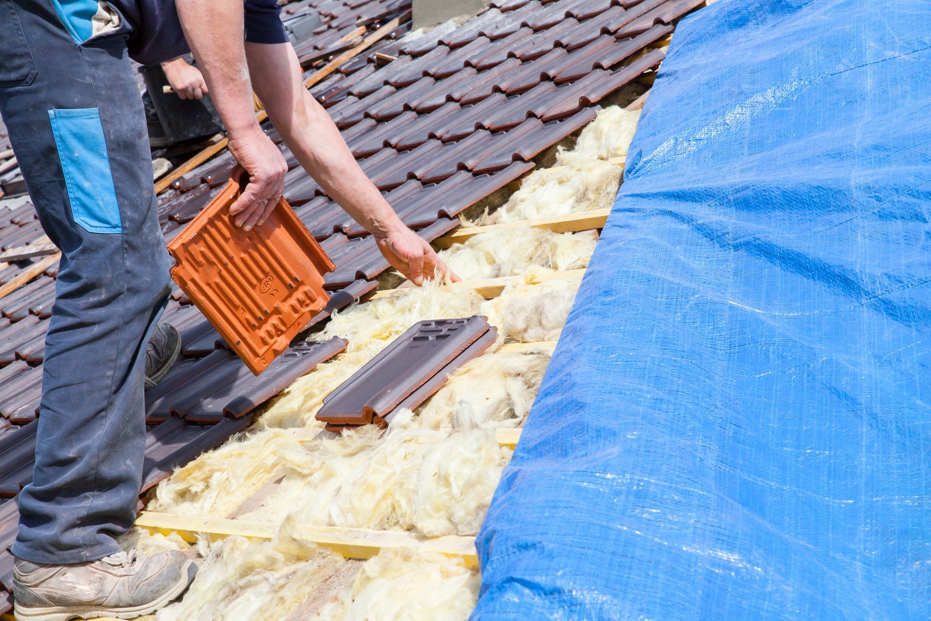 Person installing roof tiles on a roof, revealing insulation; blue tarp covers a section.