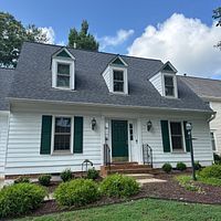White house with a dark roof and green door, featuring dormers, green shutters, and landscaping.