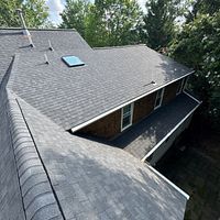 A dark gray shingled roof with a skylight on a house surrounded by green trees.