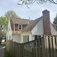 A white house with a wooden roof and large brick chimney behind a wooden fence.