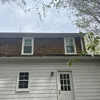 Brown shingled roof with two windows above a white-sided building with a door and a window.