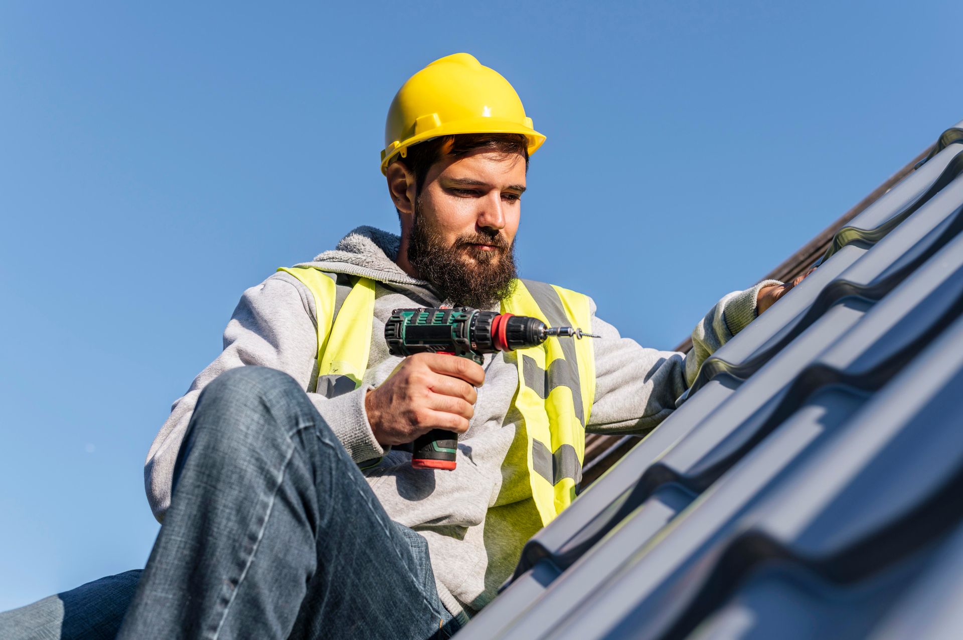 Roofer in yellow hard hat and vest, using a drill on a roof, against a blue sky.