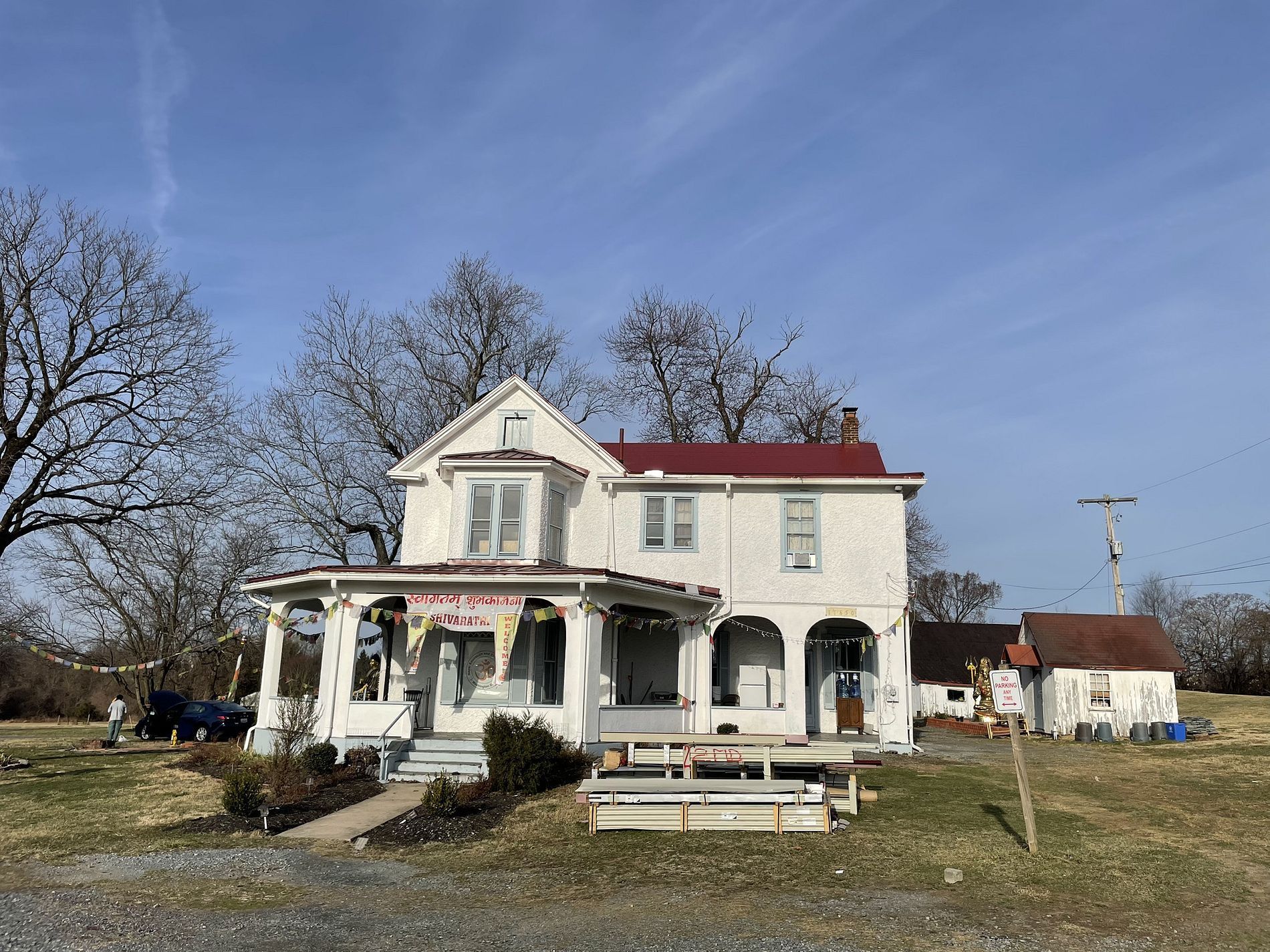 White two-story house with a red roof and porch, set on a rural property under a blue sky.