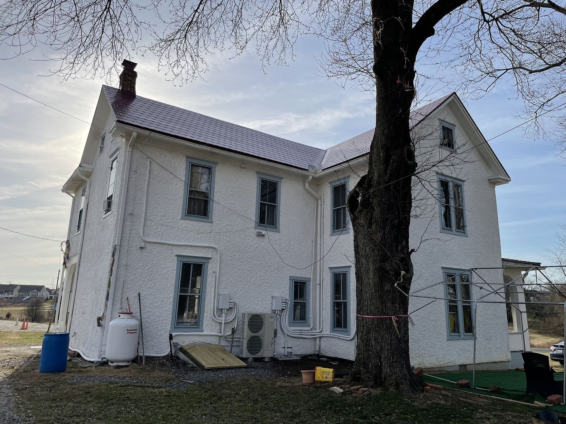 White two-story house with a dark metal roof, bare tree in the foreground. Cloudy sky.
