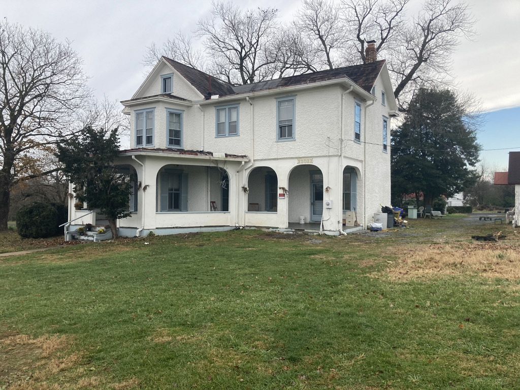 White two-story house with porch, needs repair, set in a grassy yard under a cloudy sky.