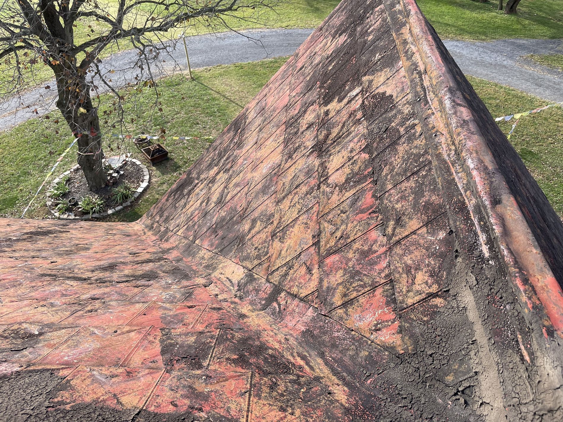 Close-up of weathered red roof with black stains; a tree and lawn are in the background.