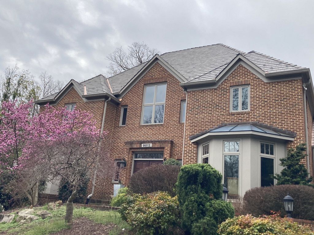 Two-story brick house with multi-toned roof; pink flowering tree and shrubs in front. Cloudy sky.