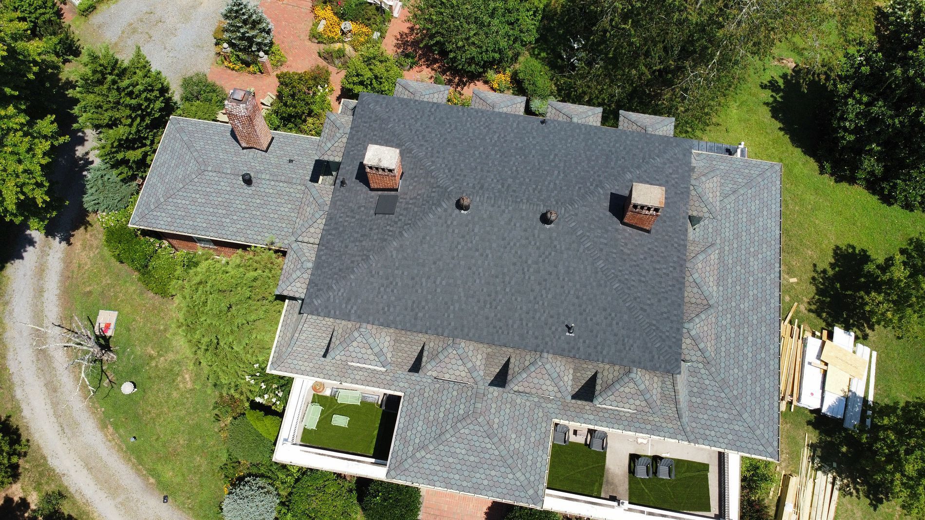 Aerial view of a dark gray roof with two chimneys, surrounded by green grass and trees.