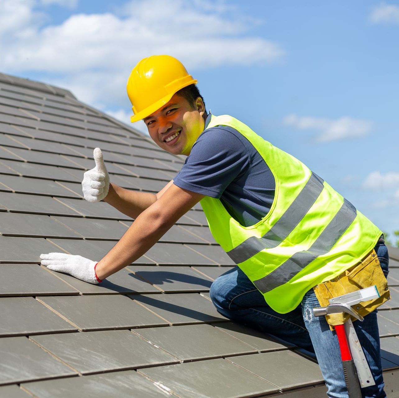 A man wearing a hard hat and safety vest is kneeling on a roof and giving a thumbs up