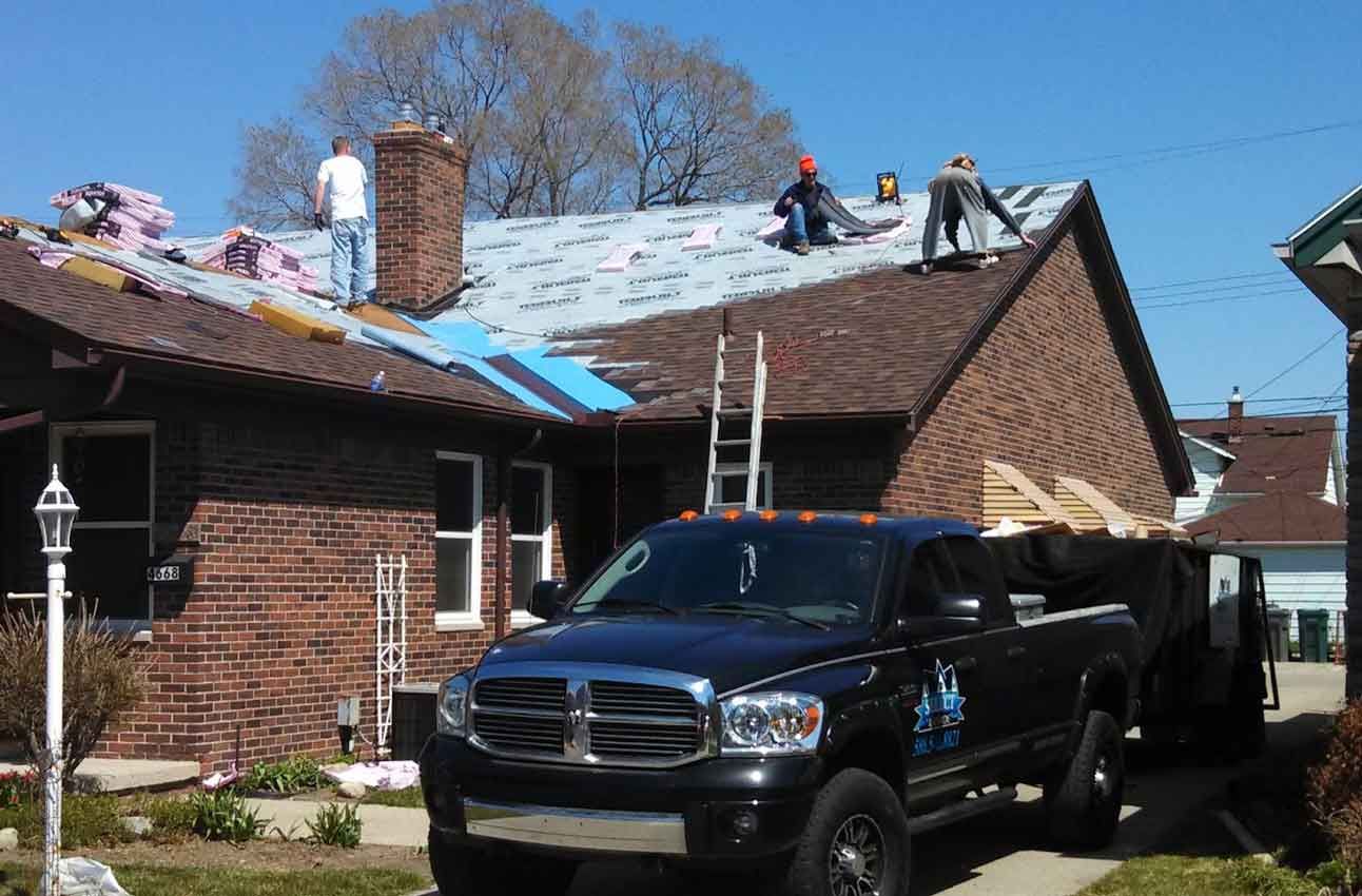 A truck is parked in front of a brick house while workers work on the roof.