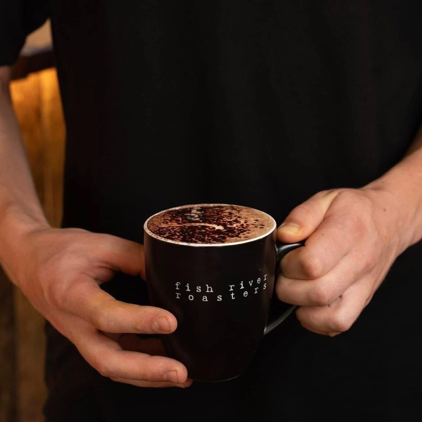 Person holding a black mug of a cappuccino, dusted with cocoa powder. — Cafe Nosh in Taree, NSW