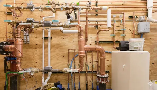 Complex copper and PVC plumbing system against a wooden wall, with pipes, valves, and a beige water tank.