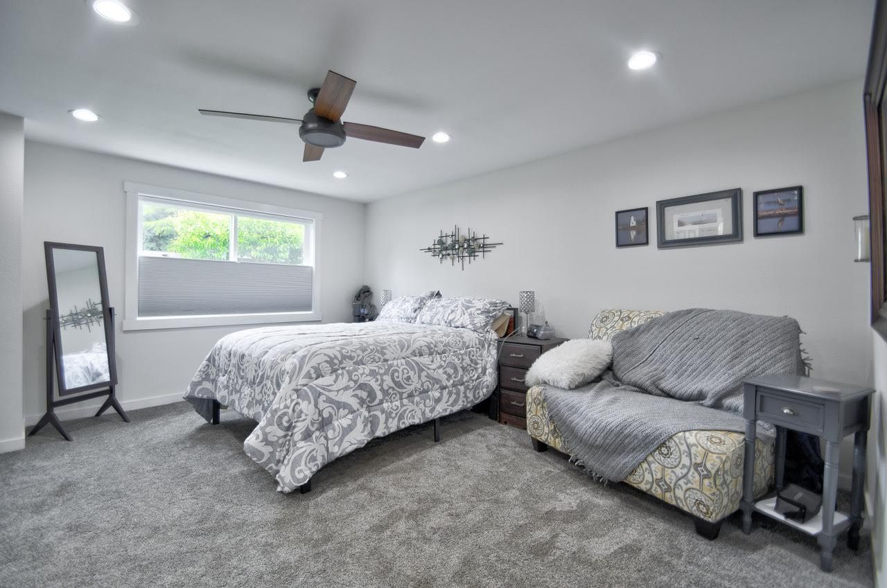 Bedroom with gray carpet, bed with patterned bedding, sofa, artwork, and a ceiling fan.