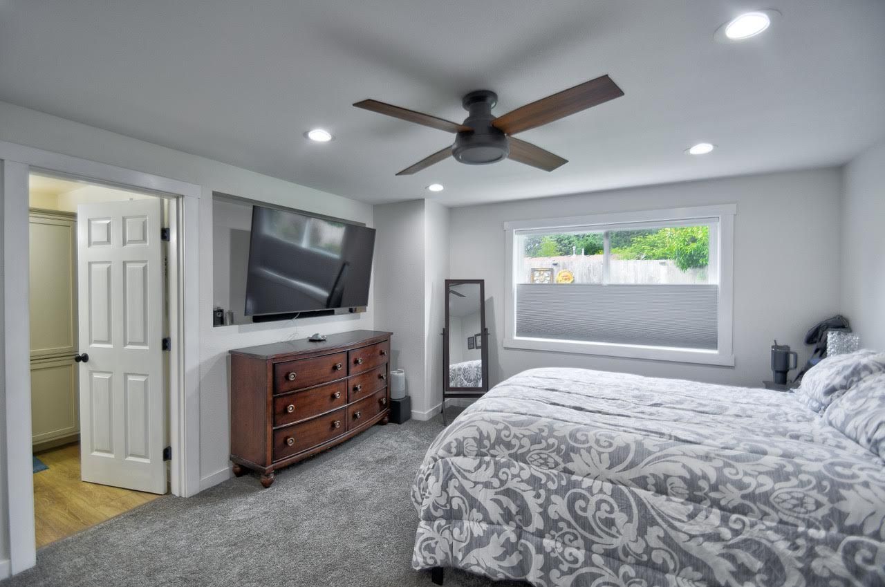 Bedroom with a bed, dresser, TV, and open doorway to a bathroom. Grey carpet, white walls, and a ceiling fan.