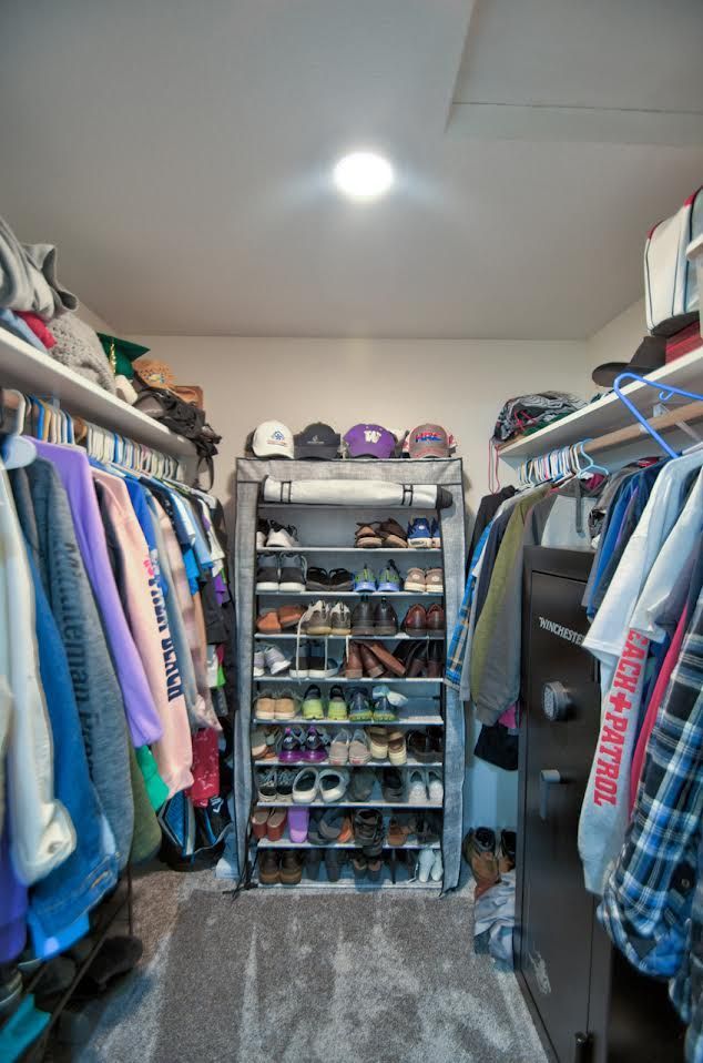 Closet with clothes on hangers and shoes on a rack. Grey carpet, white shelves, and a ceiling light.