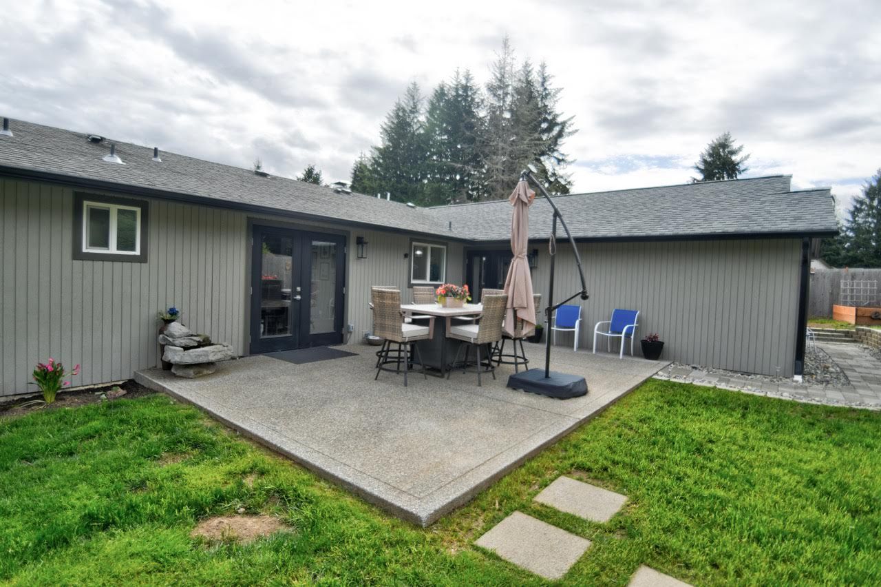 Patio with table, chairs, and umbrella outside a gray house with a green lawn and cloudy sky.