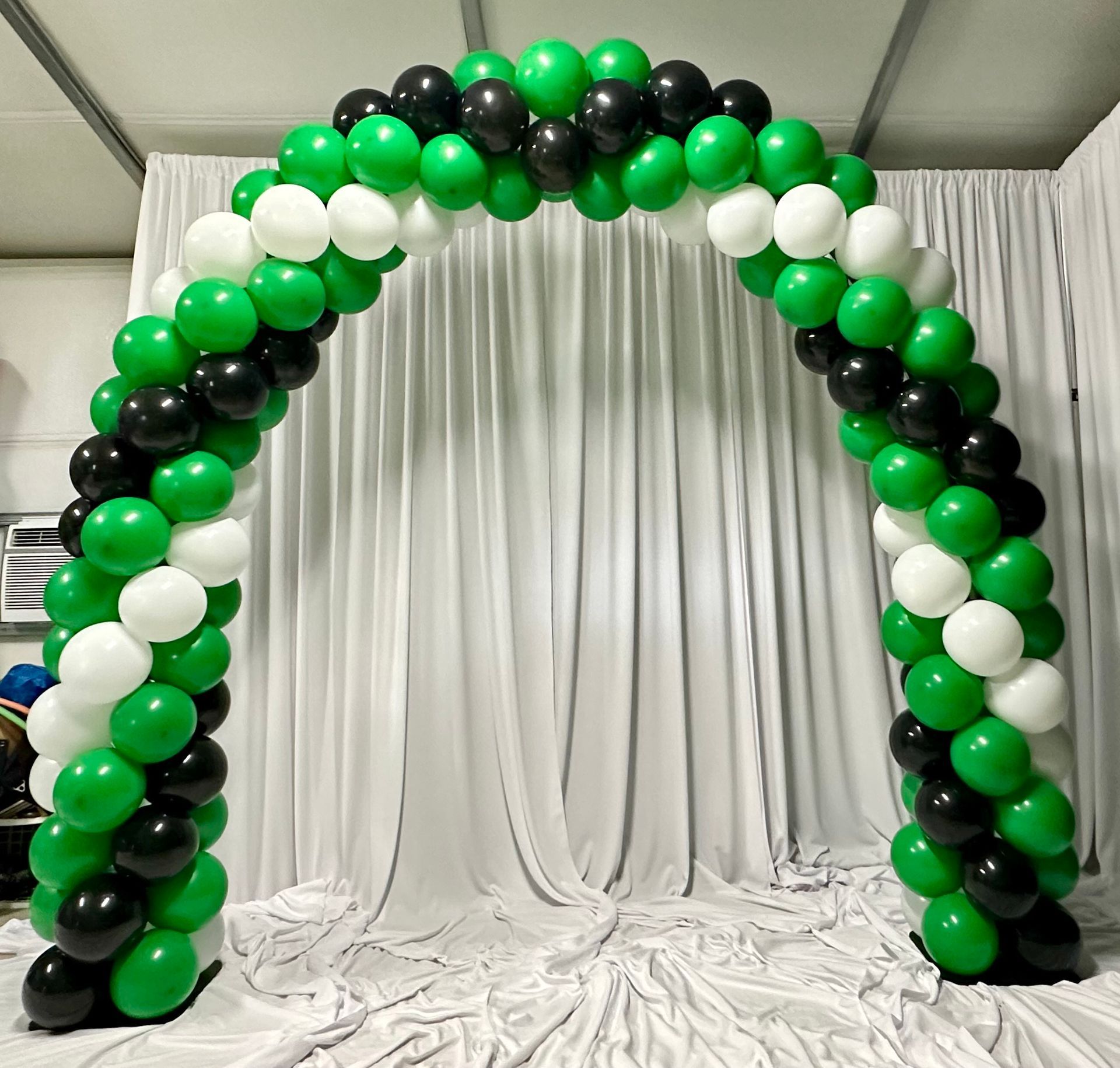 A green and black balloon arch is sitting in front of a white curtain.