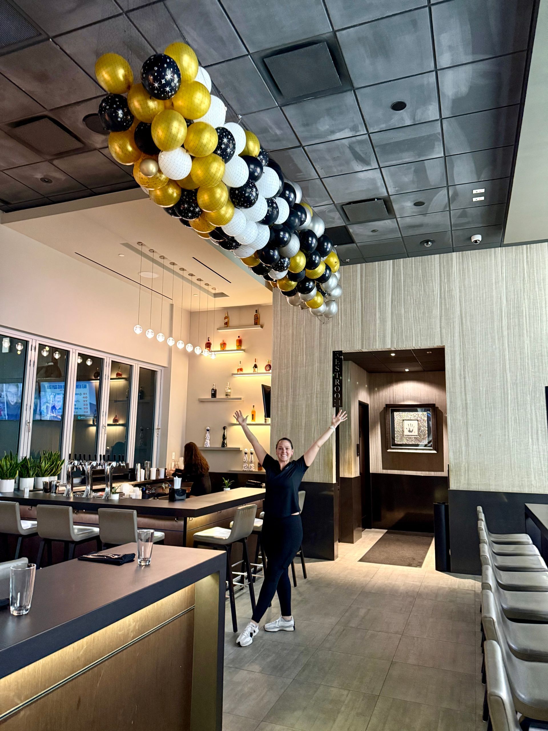 A woman is standing in a restaurant with balloons hanging from the ceiling.
