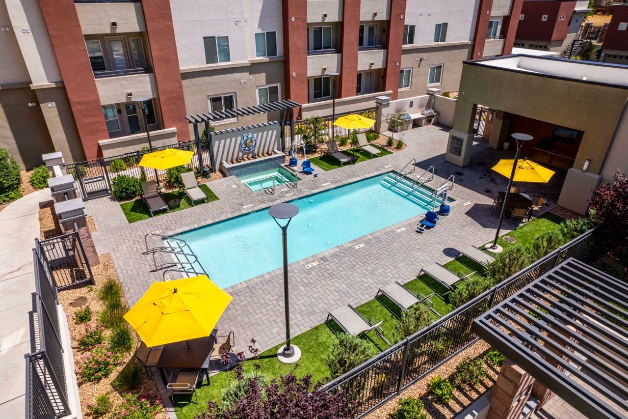 Aerial view of a rectangular pool surrounded by lounge chairs, yellow umbrellas, and apartment buildings.