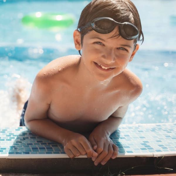 A smiling person with goggles rests their arms on the edge of a bright blue tiled swimming pool.