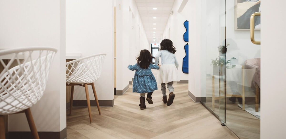 Two children walking down a bright hallway in a modern home, one in a blue dress and boots, one in a white coat.