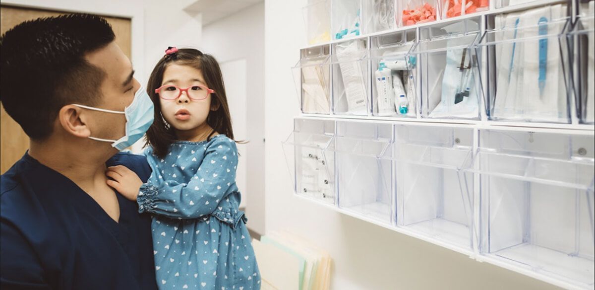 Masked adult holding child in a clinic hallway beside wall-mounted document holders