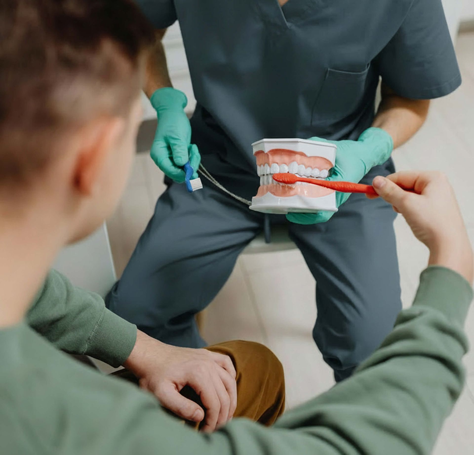 A dental professional in scrubs demonstrates proper brushing technique on a model set of teeth to a patient.