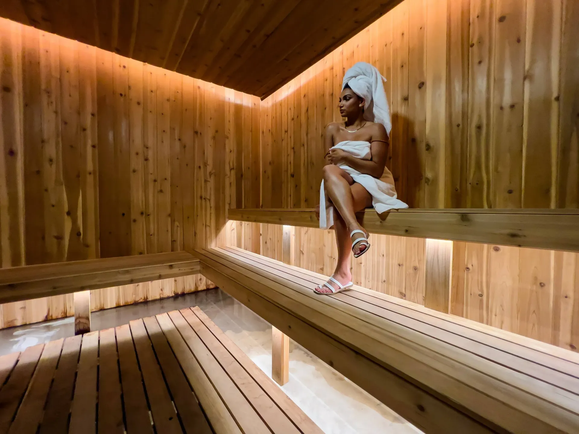 Woman seated in a wooden sauna, wrapped in a towel.