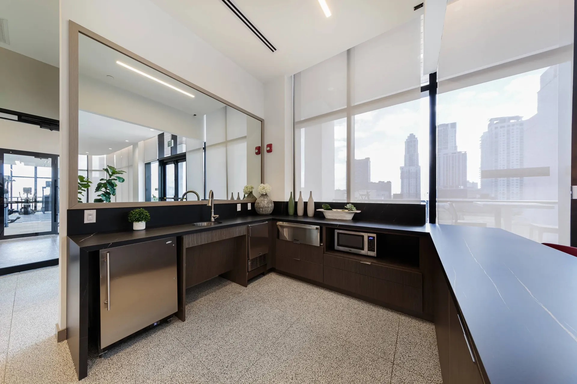 Modern communal kitchenette with dark cabinets, stainless appliances, and large windows overlooking the city.