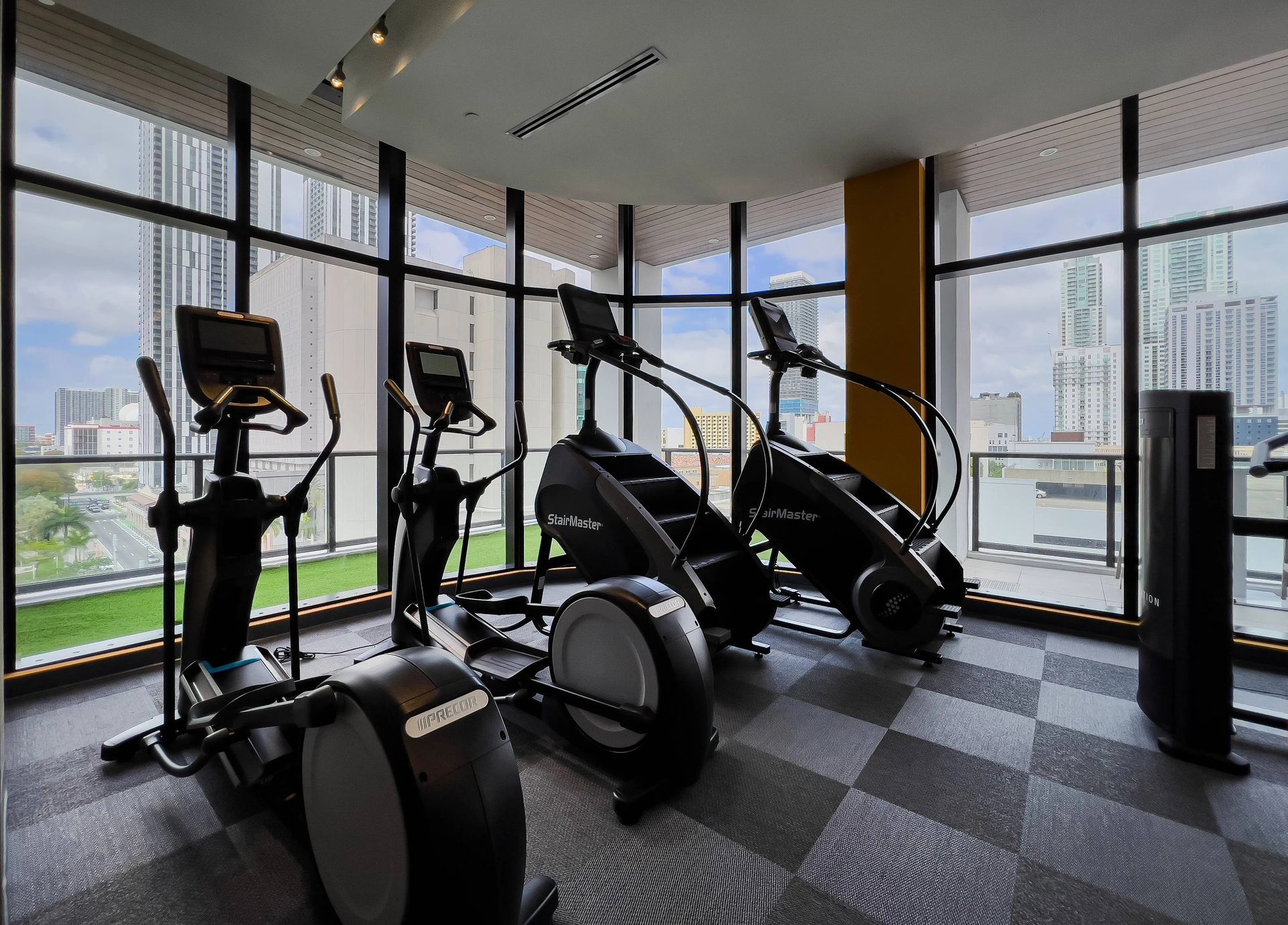 Gym with elliptical machines and stair steppers, with city views through large windows at Grand Station in Miami, FL.