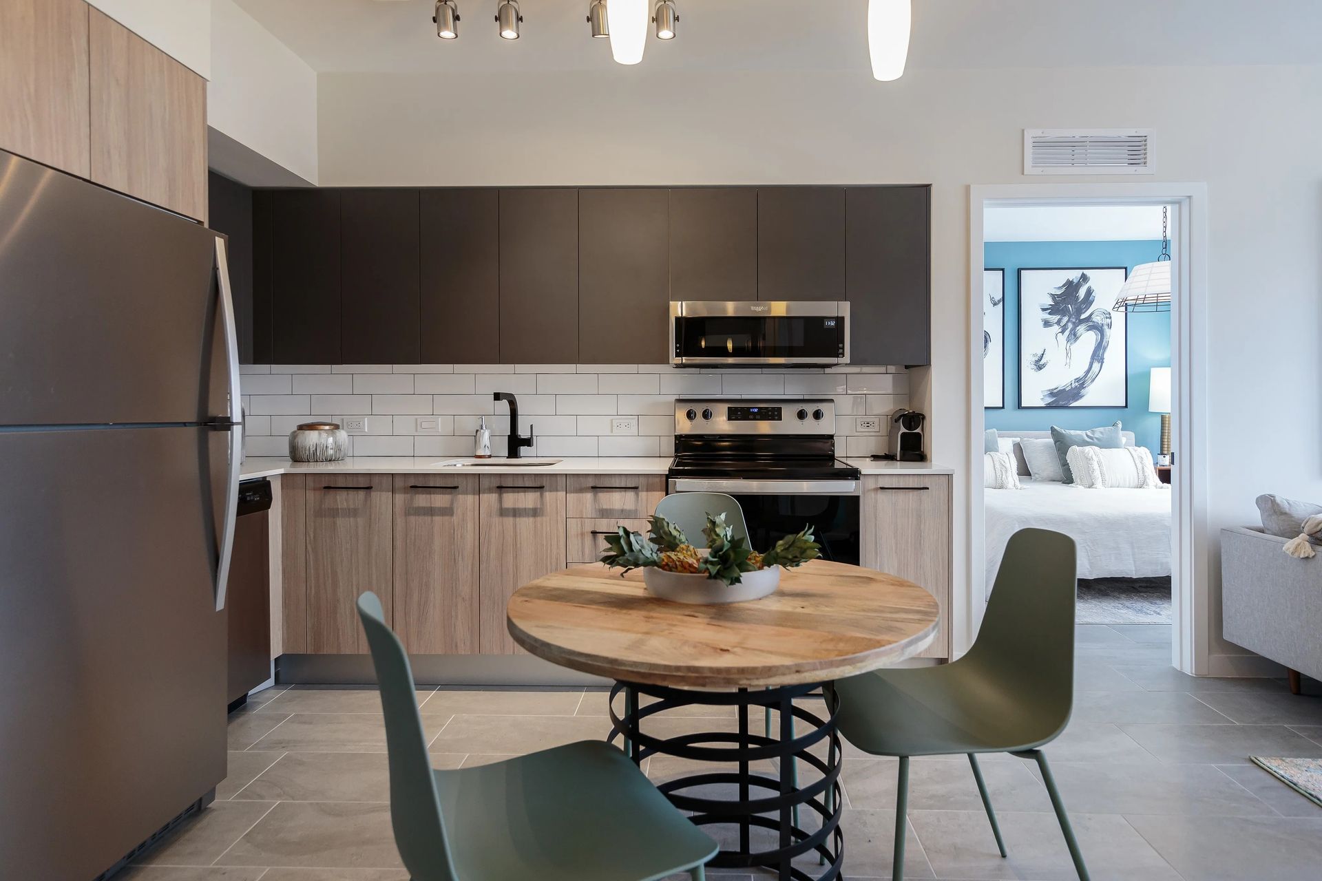 Modern kitchen with stainless steel appliances, wooden cabinets, a round dining table, and a view into a bedroom.