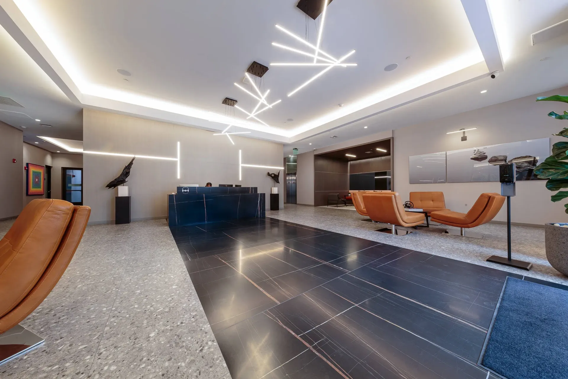 Modern apartment lobby with a reception desk, geometric hanging lights, and orange lounge chairs.