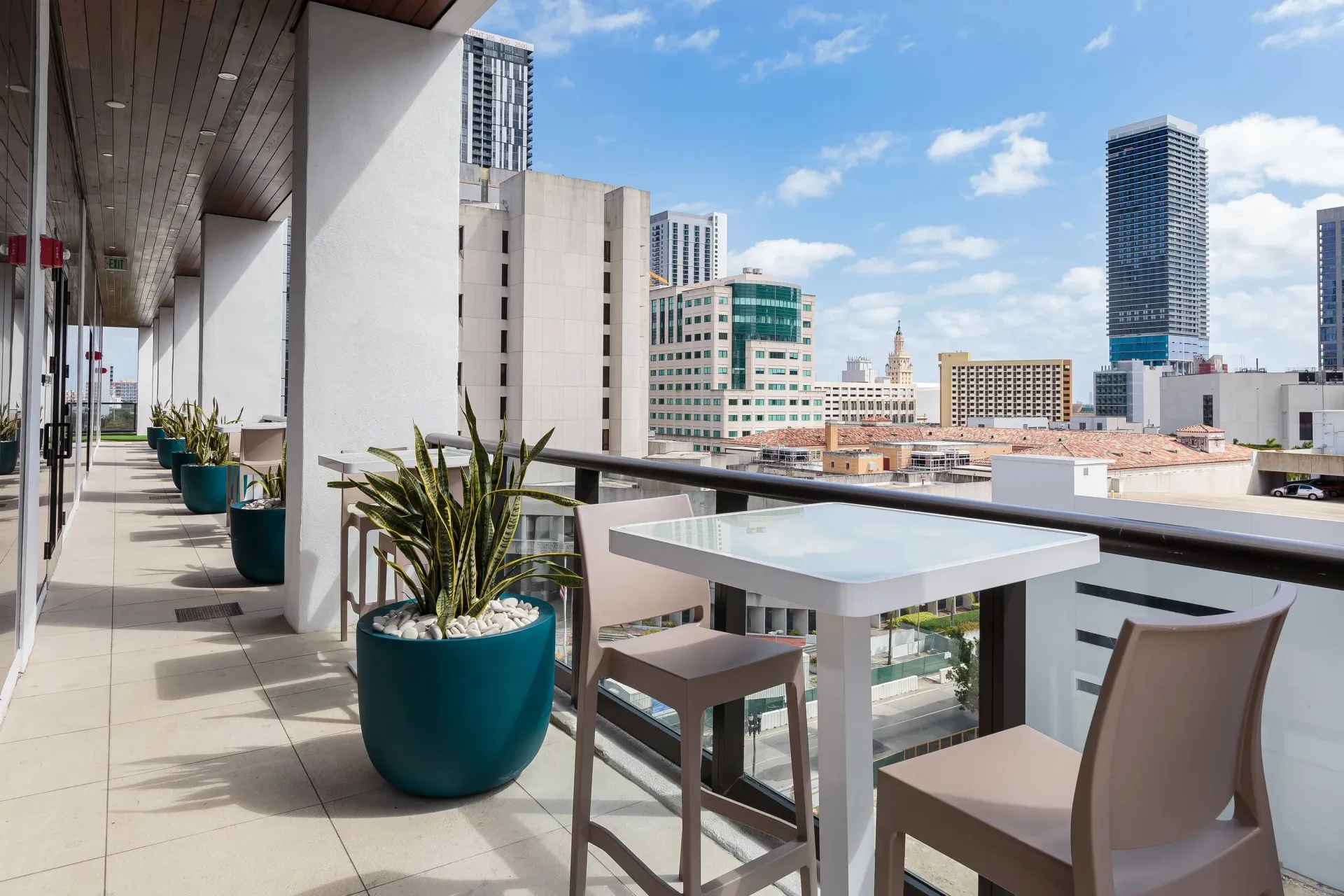 Open-air balcony with tables, chairs, and potted plants overlooking a city skyline.