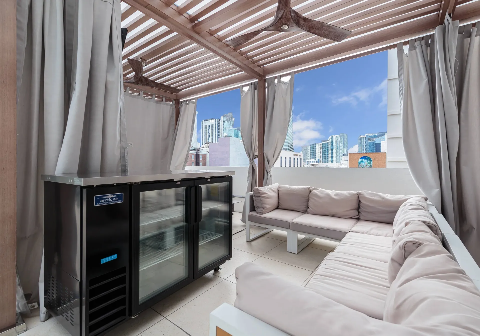 Rooftop outdoor lounge with beige sectional seating under a pergola and city skyline view.