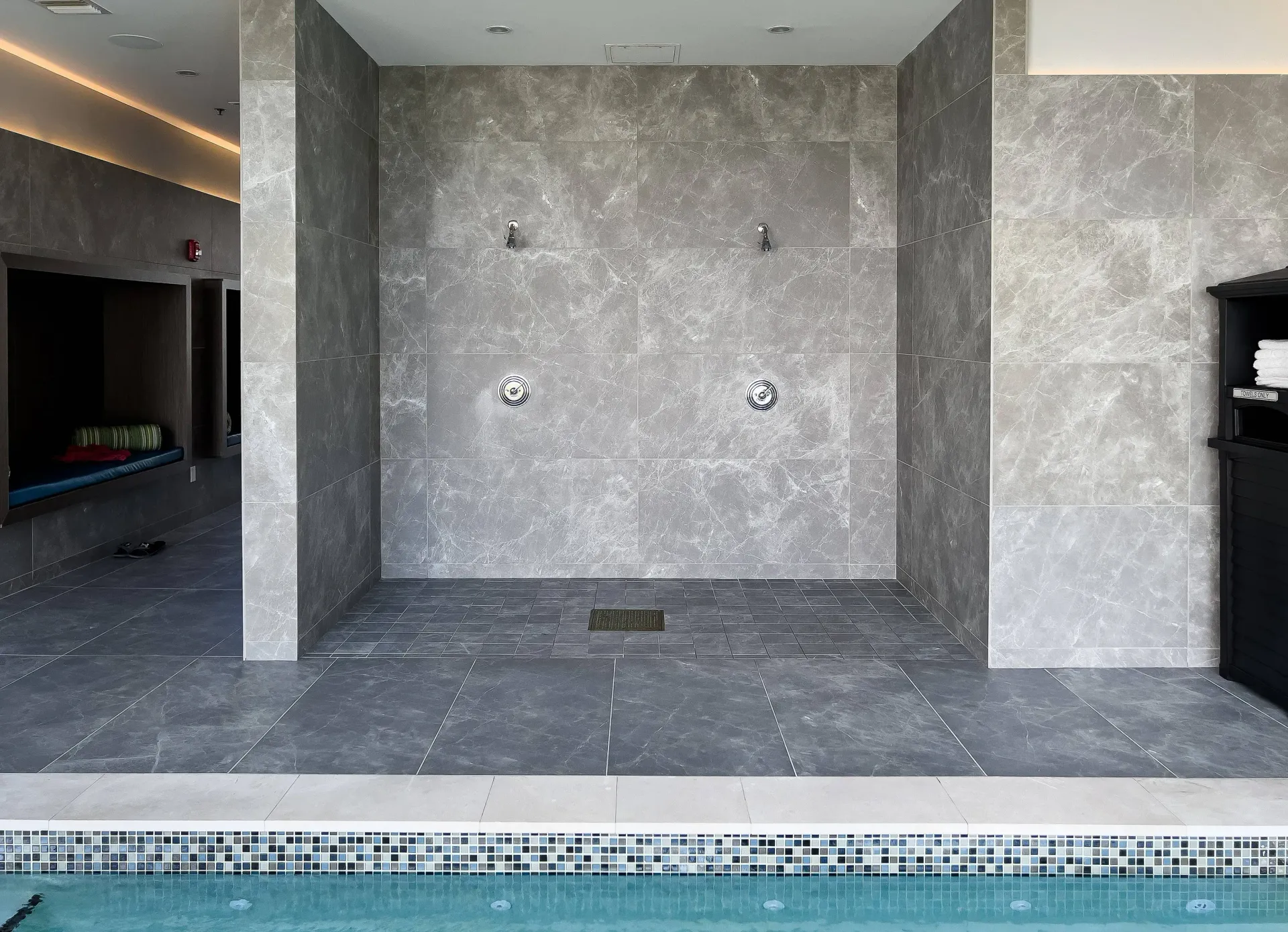 Communal pool facility shower area with gray marble tile walls and a mosaic pool edge.