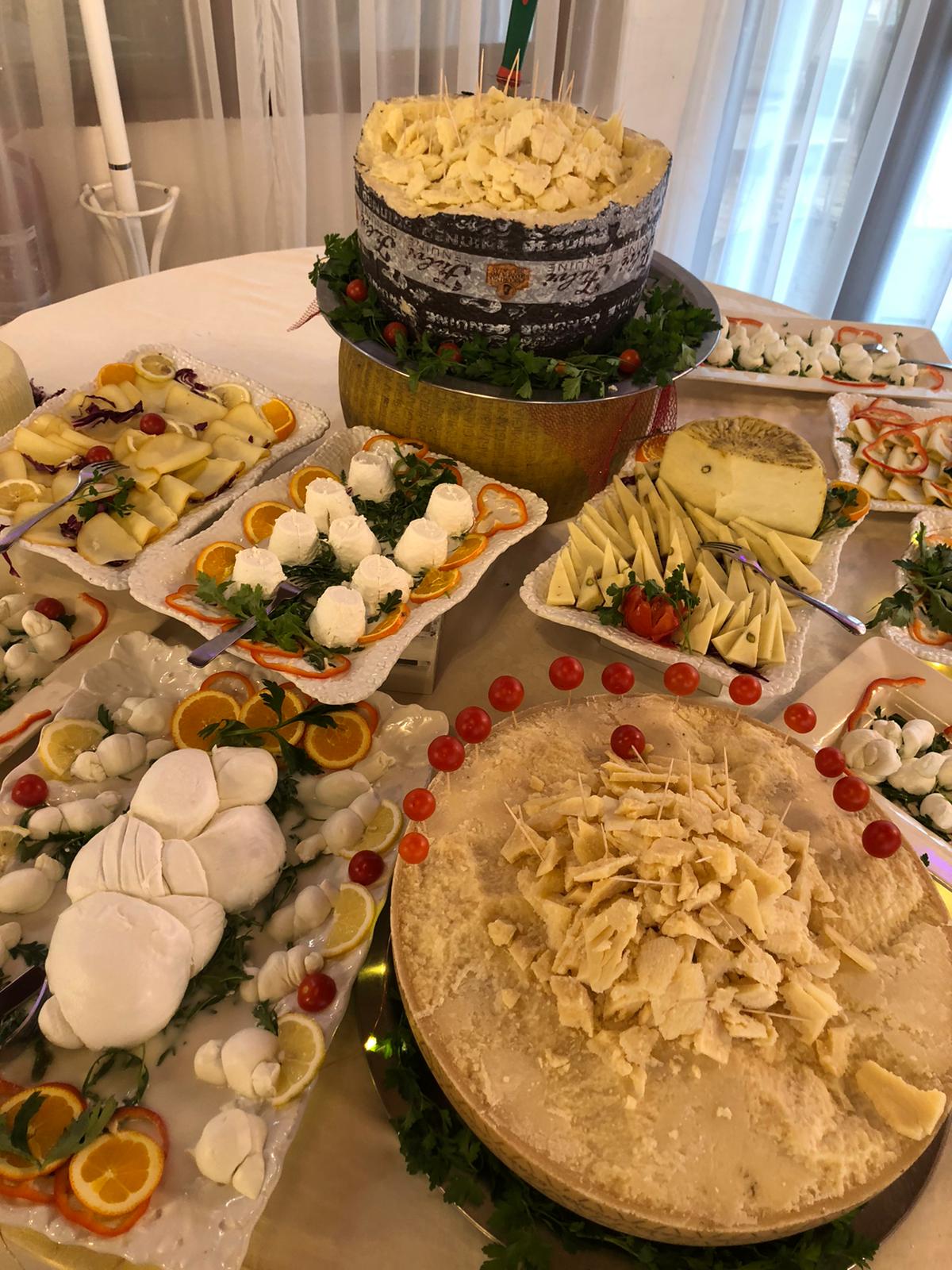 Una tavola imbandita con vari formaggi, tra cui una grande forma di formaggio e una torta a strati, guarnita con verdure e fette d'arancia.