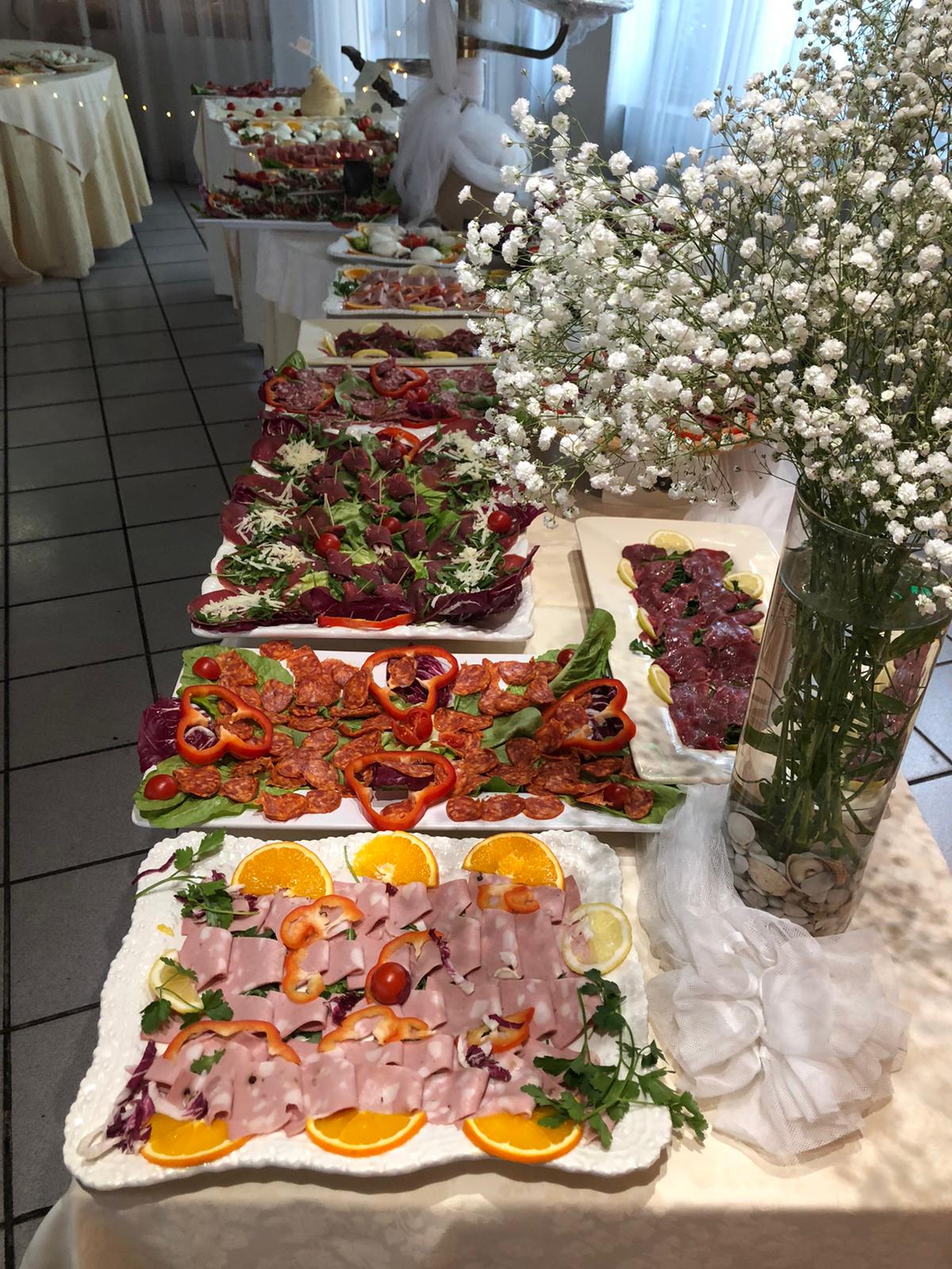 Tavolo da buffet con vari piatti. Include salumi, verdure e frutta, con un vaso di fiori bianchi.